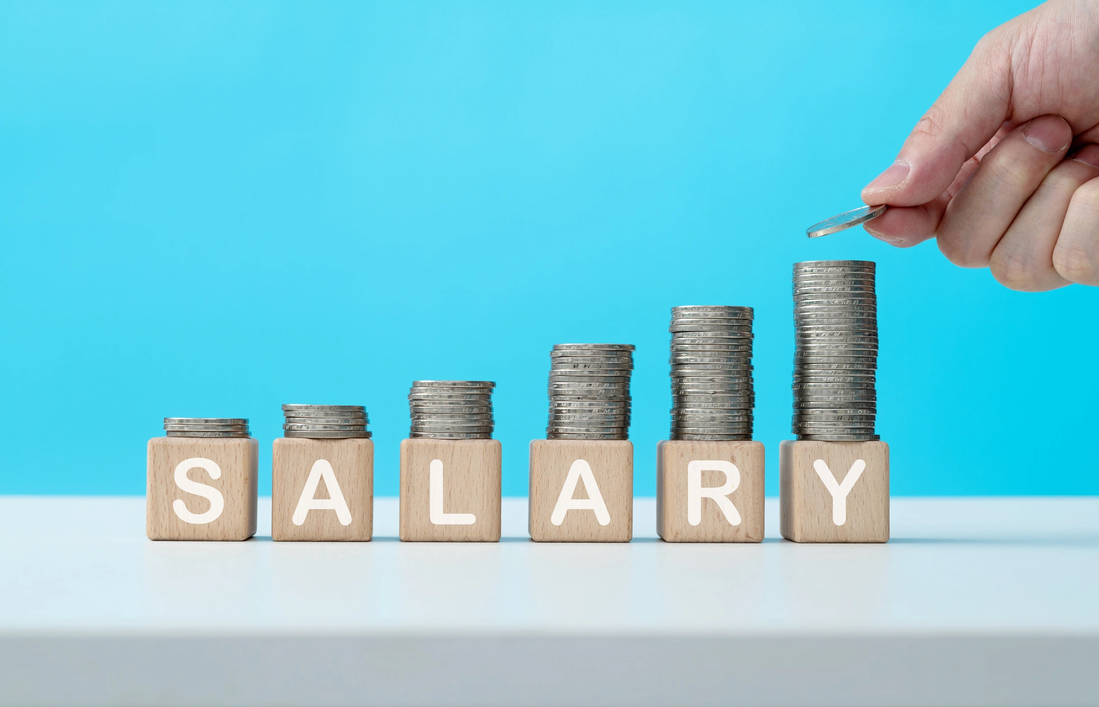 Multiple stacks of coins, going from a smaller pile, to a larger pile, with the word 'salary' spelt out in blocks in front of the stacks of coins - this depicts rising costs due to the 2026 National Minimum Wage rise