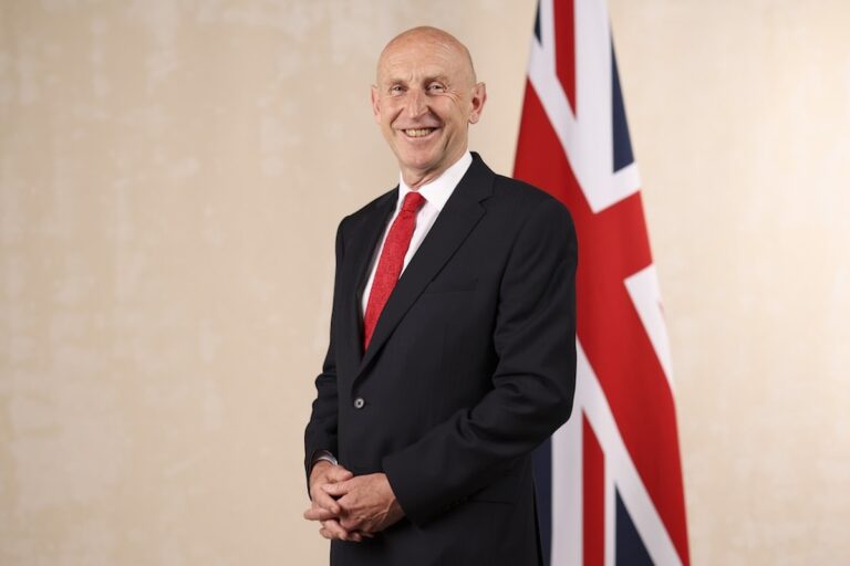 The official portrait of Chancellor John Healy, posed in front of a beige wall with a union jack flag behind him